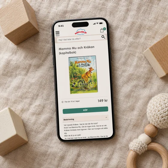 Babyland's product page for the chapter book Mamma Mu och Kråkan, with its cover, price and description, on a phone lying on a linen blanket among wooden toy blocks.