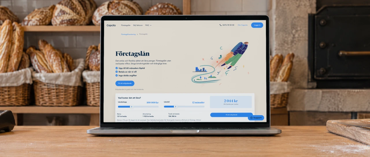 Capcito's page “Företagslån” (business loans) open on a laptop on the oak counter of a small bakery in the morning, with sourdough loaves and baguettes in baskets on the shelves, a stone oven and a card terminal.