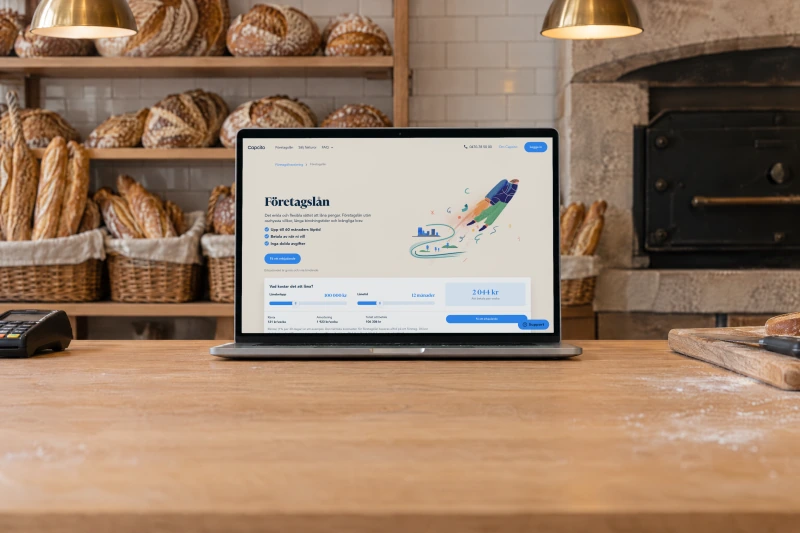 Capcito's page “Företagslån” (business loans) open on a laptop on the oak counter of a small bakery in the morning, with sourdough loaves and baguettes in baskets on the shelves, a stone oven and a card terminal