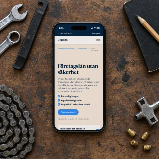 Capcito's page “Företagslån utan säkerhet” (loans without collateral) on a phone lying on a worn workbench in a bicycle repair shop, beside spanners, a chain, a notebook and brass valve caps.