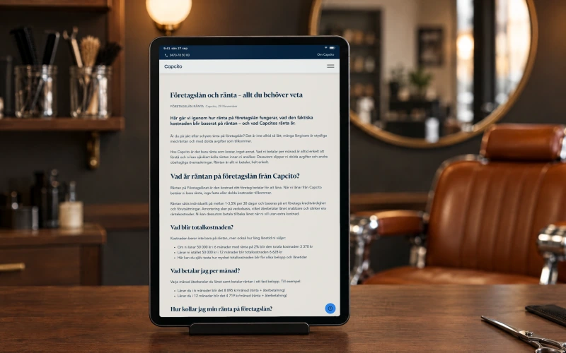 Capcito's article “Företagslån och ränta - allt du behöver veta” on a tablet in a stand on a barbershop's reception counter, with a leather barber chair, a round mirror and globe lights behind.