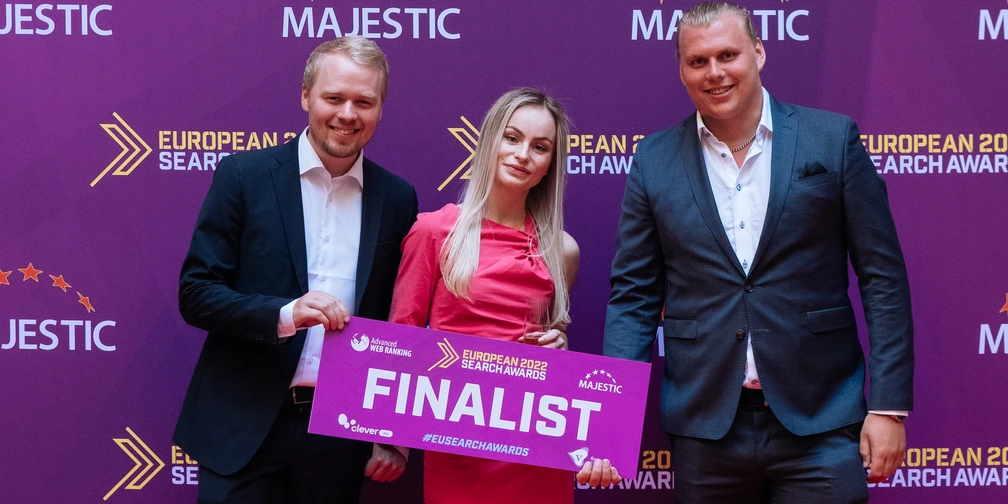 Stella Wannberg holding a European Search Awards 2022 finalist sign between two men in suits, in front of the sponsor wall.