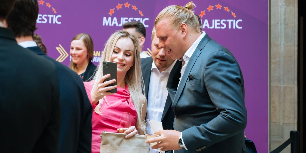 Stella Wannberg smiling at her phone, a glass in her hand, beside two men in suits at the sponsor wall.
