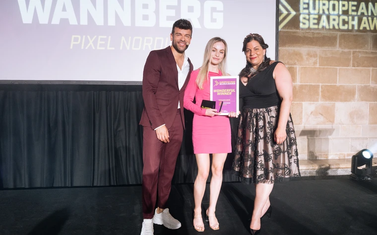 Stella Wannberg in a pink dress on stage holding the winner's diploma, between the host and the award presenter, with her name and Pixel Nordic on the screen behind them.