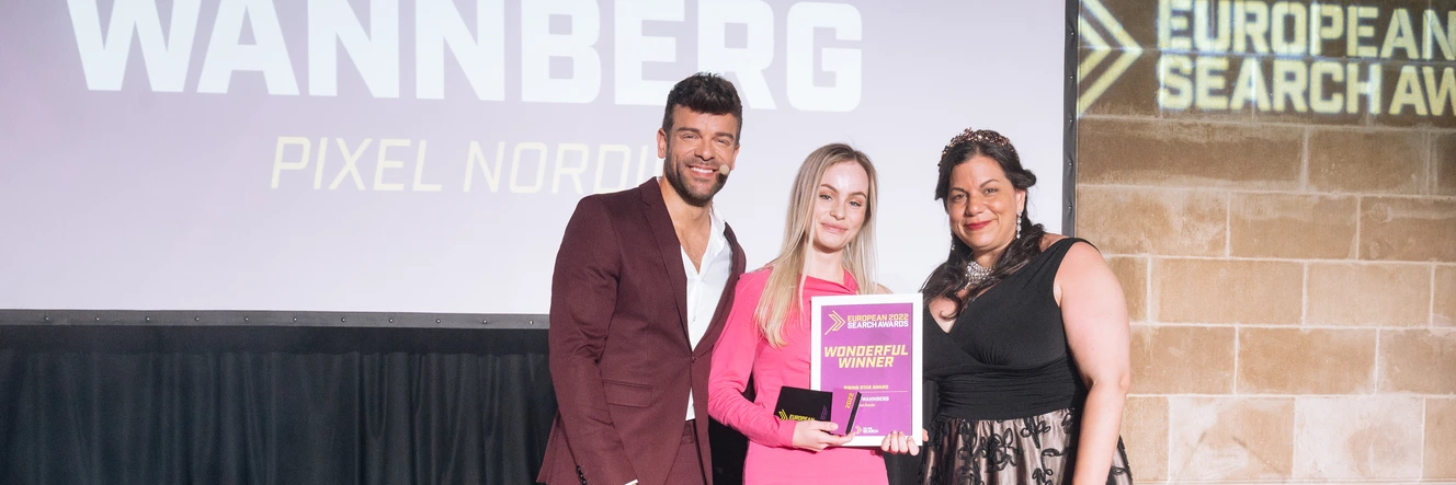 Stella Wannberg in a pink dress on stage holding the winner's diploma, between the host and the award presenter, with her name and Pixel Nordic on the screen behind them.