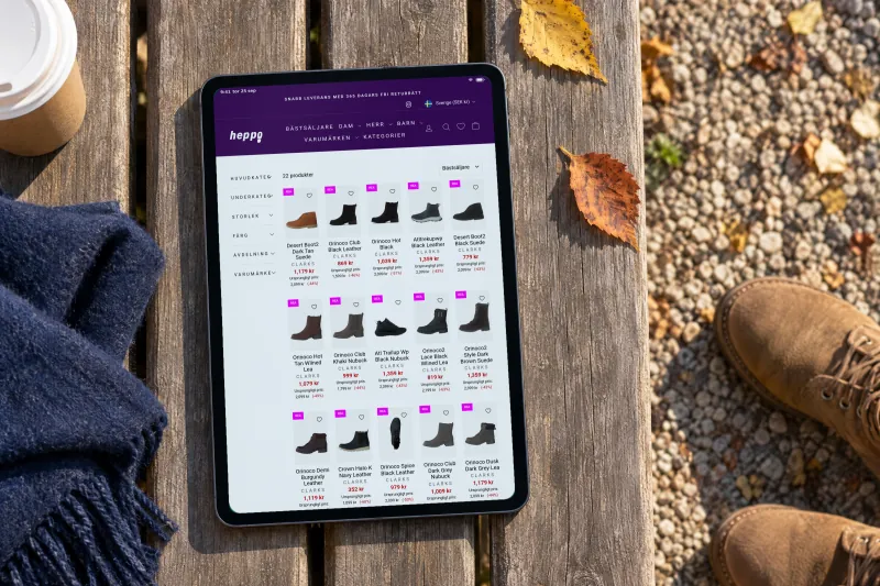 Heppo's page of Clarks boots, with a grid of boots and their prices, on a tablet lying on a wooden park bench in low autumn sun, beside a navy scarf, a takeaway cup and a pair of tan suede boots on the gravel