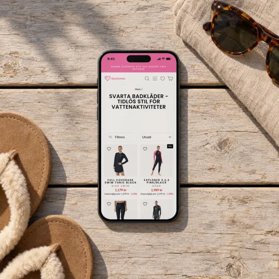 Sportamore's page for black swimwear, with its title and swimsuits below, on a phone lying on a sunlit wooden deck beside flip-flops and sunglasses.