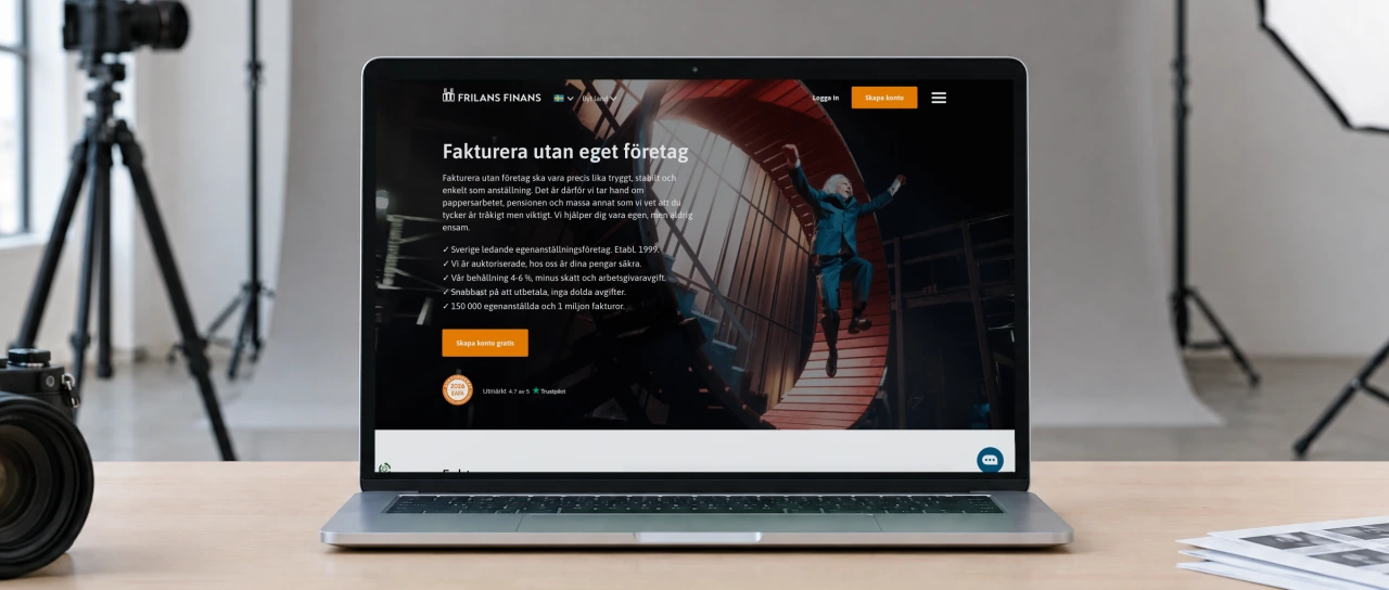 Frilans Finans' Swedish home page “Fakturera utan eget företag” (invoicing without your own company) on a laptop on a birch table in a photographer's studio, with grey backdrop paper, a softbox and a camera on a tripod behind.