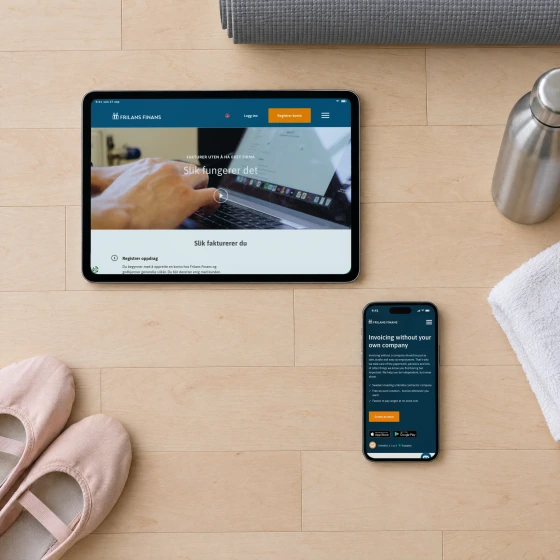 A tablet lying sideways and a phone on the maple floor of a dance studio, showing Frilans Finans' Norwegian page “Slik fungerer det” and the English page “Invoicing without your own company”, beside ballet shoes, an exercise mat and a water bottle.
