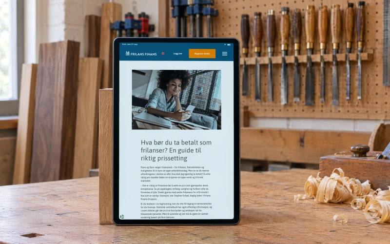 Frilans Finans' Norwegian guide “Hva bør du ta betalt som frilanser?” (what should you charge as a freelancer) on a tablet on a beech workbench in a woodworking shop, beside a hand plane and wood shavings.