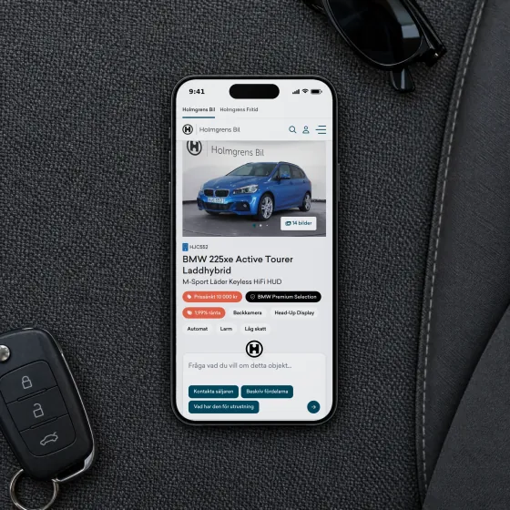 Holmgrens Bil's page for a BMW 225xe Active Tourer plug-in hybrid, with its photo, equipment and a field for questions, on a phone lying on a car's passenger seat beside a car key and sunglasses.