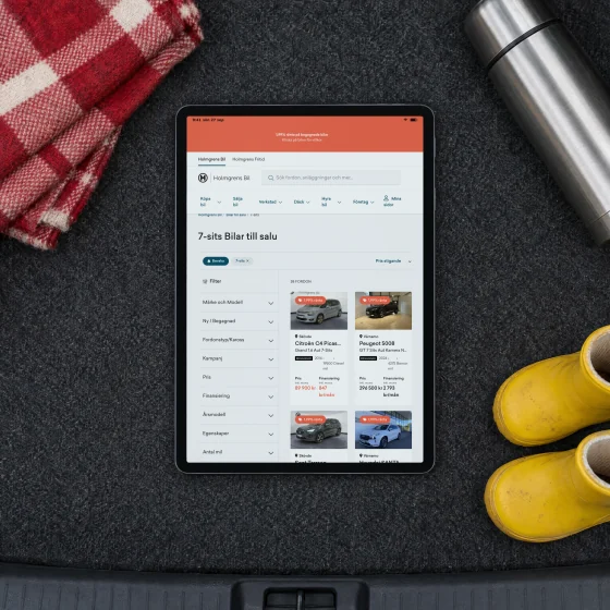 Holmgrens Bil's page for seven-seat cars for sale on a tablet lying in the boot of an estate car, beside a checked picnic blanket, a thermos and a pair of yellow children's boots.