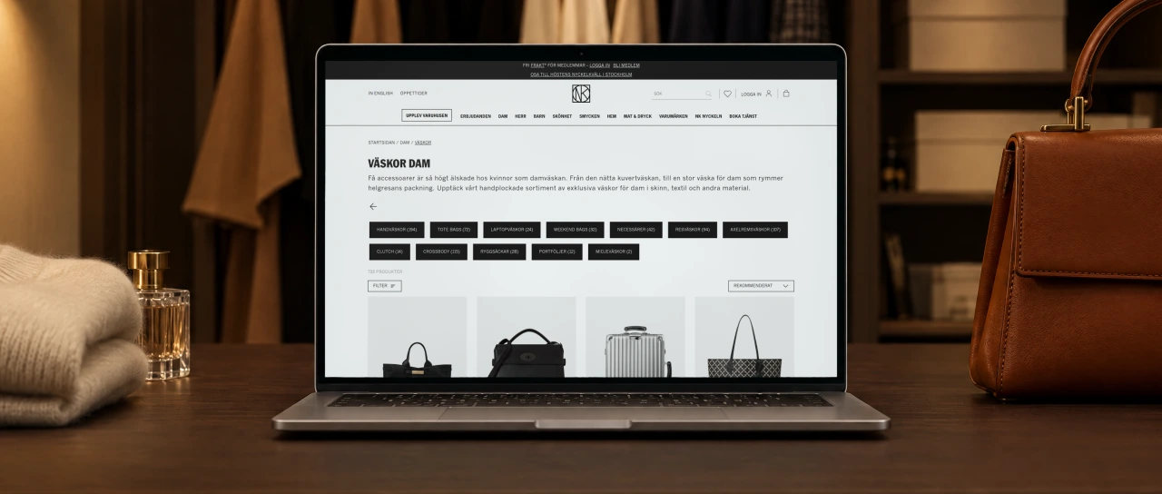 NK's category page for women's bags, with its title, a short intro, a row of subcategories and the first bags, open on a laptop on a walnut dressing table in a walk-in wardrobe in the evening, beside a cognac leather handbag and a folded cashmere sweater.