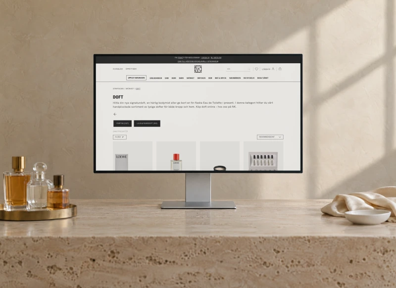 NK's fragrance category page, with its title, intro and the first perfumes, on a desktop monitor on a travertine desk against a limewashed wall, with perfume bottles on a brass tray.