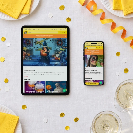 A tablet and a phone on a table laid with a white linen cloth and yellow napkins, showing Partyking's Danish page “Halloweenpynt” and Finnish page “Halloween Meikki”.