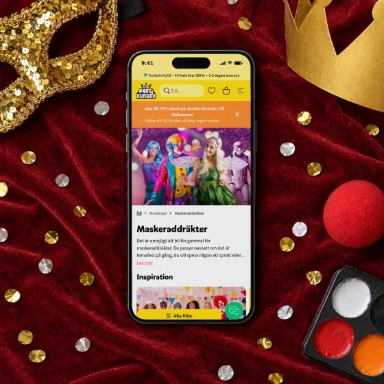 Partykungen's page “Maskeraddräkter” on a phone lying on a red velvet cape, beside a gold sequin mask, a paper crown, a red clown nose, a face paint palette and confetti.