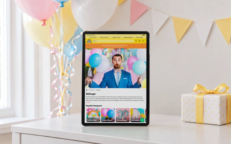 Partykungen's page “Ballonger” on a tablet standing on a white sideboard in a room decorated for a birthday, with pastel balloons, bunting, a wrapped gift and paper cups.