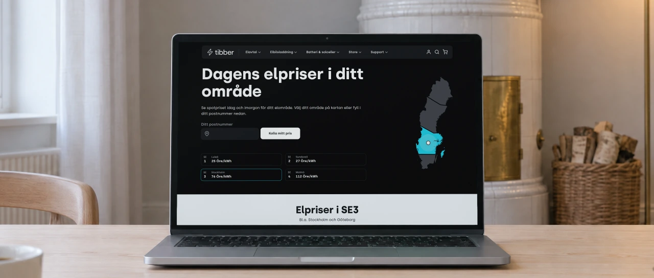 Tibber's page “Dagens elpriser i ditt område”, today's electricity prices in Sweden's four price areas beside a map, open on a laptop on a birch dining table in a living room with a white tiled stove and a basket of firewood behind.