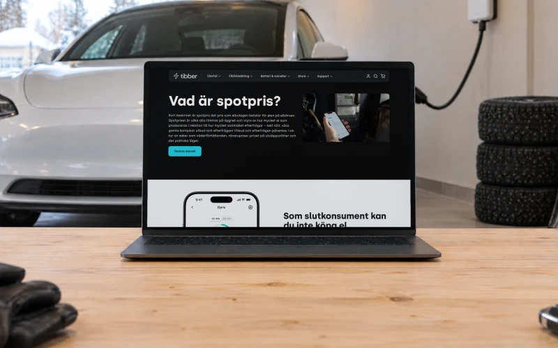 Tibber's page “Vad är spotpris?”, explaining the spot price, on a laptop on a workbench in a home garage, with an electric car plugged into a wall charger behind it.