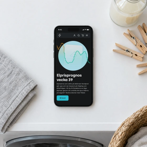 Tibber's electricity price forecast for week 39 on a phone lying on top of a washing machine, beside a folded towel, wooden clothes pegs and a laundry basket.
