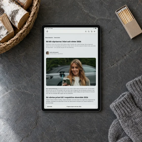 Tibber Magazine's article “Så blir elpriserna i höst och vinter 2026” on a tablet lying on a slate hearth, beside a basket of birch logs, a pair of wool socks and a box of matches.