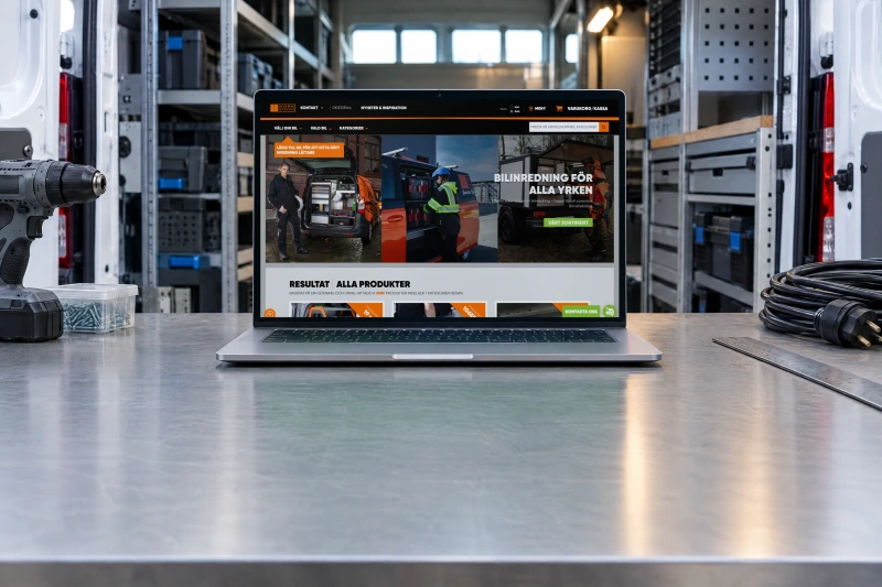Work System's home page, with its car selector and product categories, open on a laptop on a steel workbench in a van outfitting workshop, in front of a white van with its rear doors open and shelving fitted inside