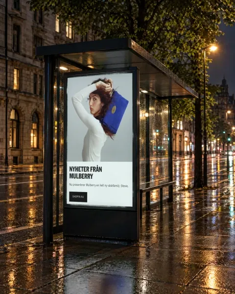 The NK homepage campaign on a lit poster in a bus shelter, on a rainy street at night