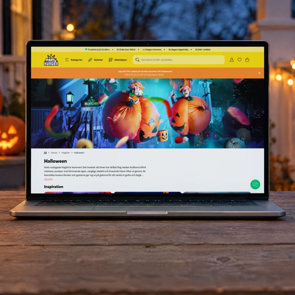 Partykungen's Halloween page open on a laptop on a weathered wooden table on a porch at dusk, between glowing carved pumpkins, a bowl of sweets and a string of warm lights along the railing