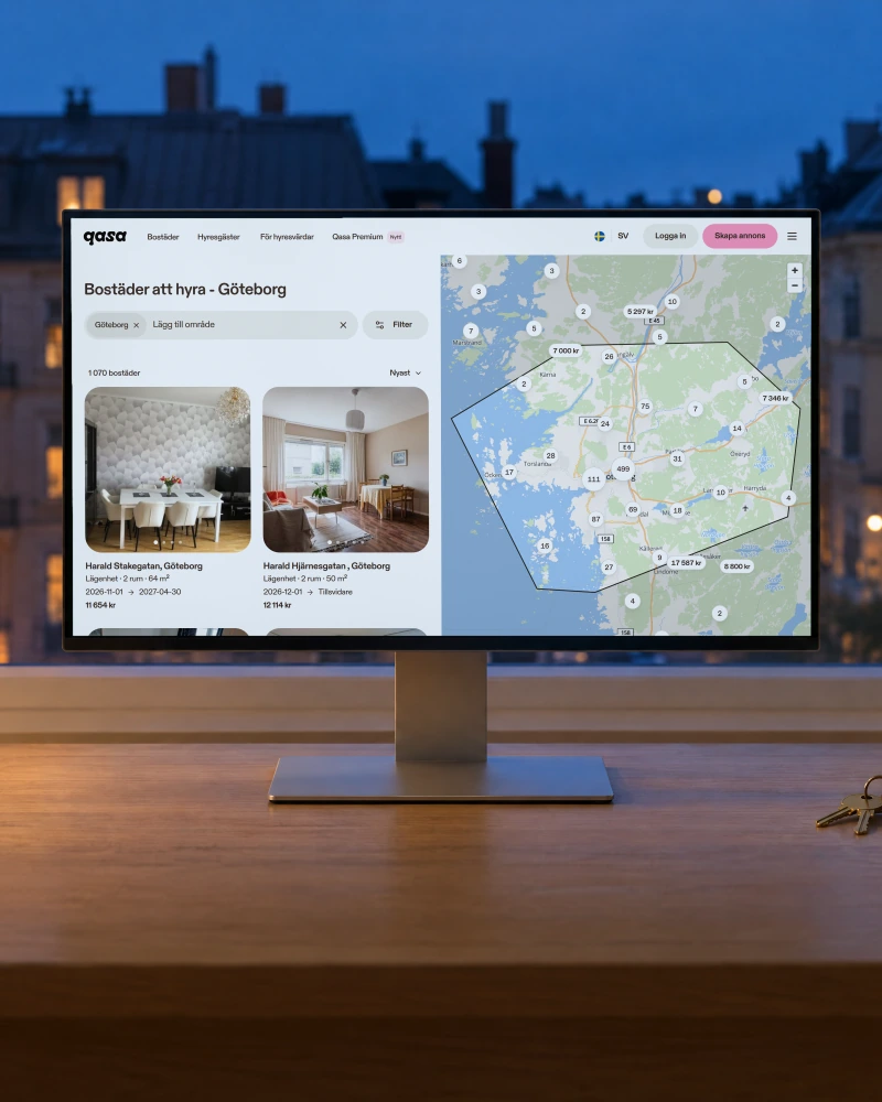 Qasa's page of homes to rent in Gothenburg, with a map of listings and two home cards, on a desktop monitor on an oak desk by a window at dusk, with a table lamp, a mug and a set of keys
