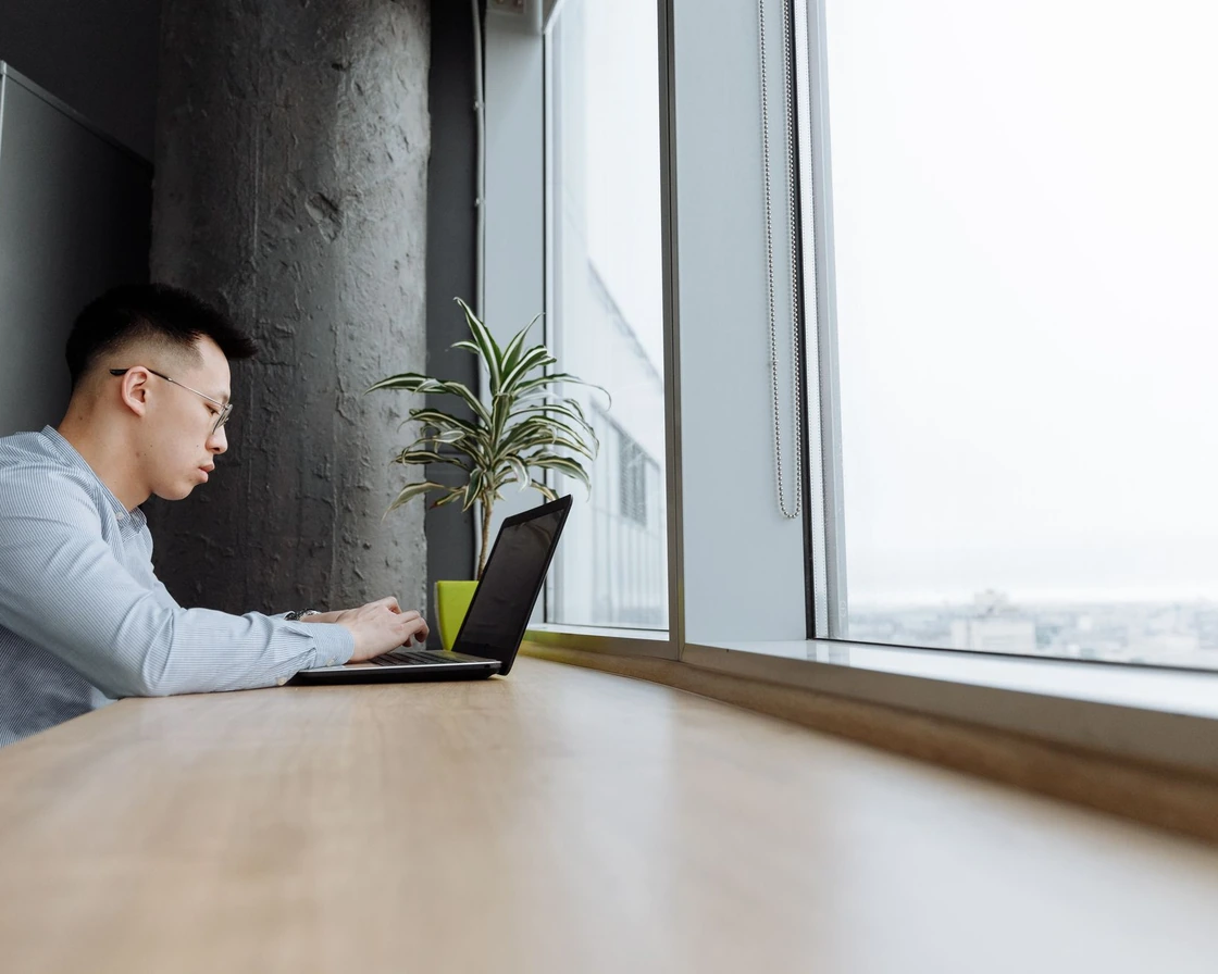 A person working on a laptop at a long desk by a window