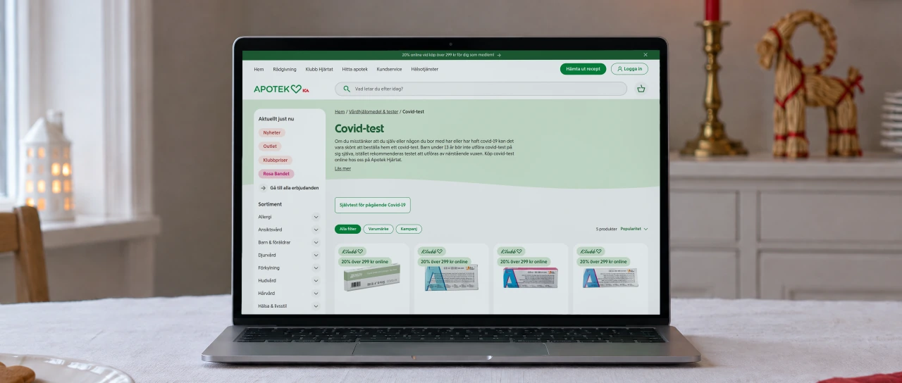 Apotek Hjärtat's category page “Covid-test”, with its introduction and the first self-tests, open on a laptop on a table with a white linen cloth, beside gingerbread biscuits and a red napkin, with a lit red candle and a straw goat behind.