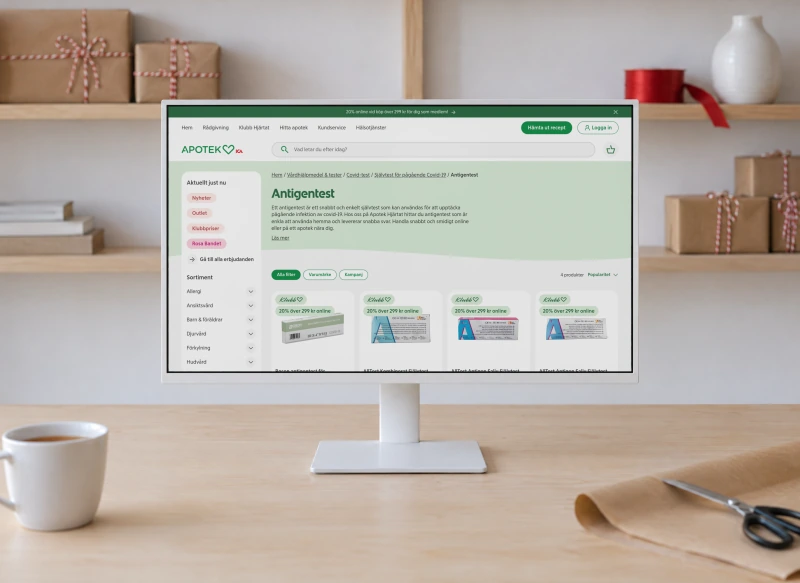 Apotek Hjärtat's page “Antigentest” on a white monitor on a birch desk, in front of shelves of presents wrapped in brown paper with red and white string.