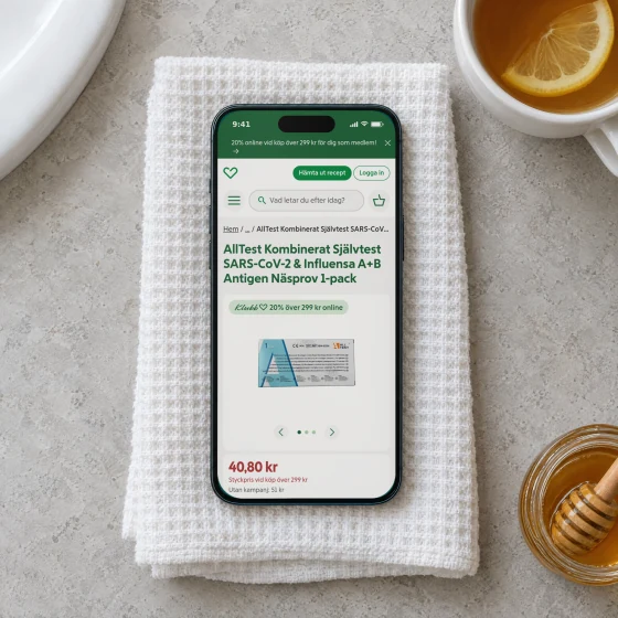 Apotek Hjärtat's product page for a combined self-test for covid-19 and influenza on a phone lying on a folded white towel on a bathroom counter, beside a mug of tea with lemon and a jar of honey.