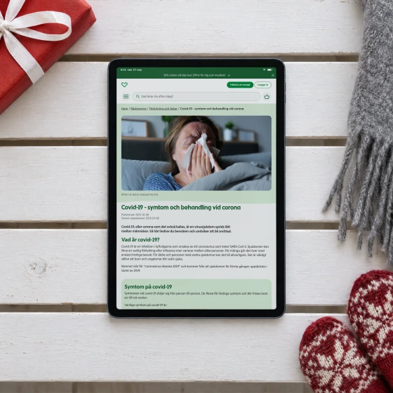 Apotek Hjärtat's advice page “Covid-19 - symtom och behandling vid corona” on a tablet lying on a white hallway bench, beside a red present, a grey scarf and red and white mittens.