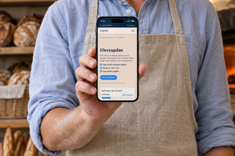 Capcito's page “Företagslån” (business loans) on a phone held up by a baker in a linen apron, with sourdough loaves on the shelves and a stone oven behind