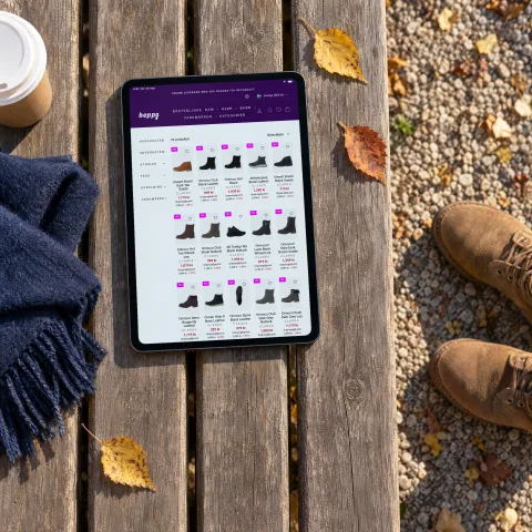 Heppo's page of Clarks boots, with a grid of boots and their prices, on a tablet lying on a wooden park bench in low autumn sun, beside a navy scarf, a takeaway cup and a pair of tan suede boots on the gravel
