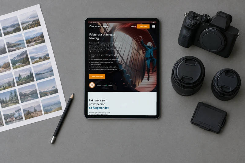 Frilans Finans' Swedish home page “Fakturera utan eget företag” (invoicing without your own company) on a tablet lying on a grey work table in a photographer's studio, beside a camera, two lenses and a printed contact sheet