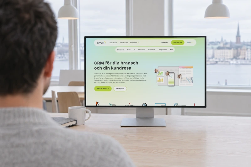 Lime Technologies' page for Lime CRM, CRM för din bransch och din kundresa, on a desktop monitor seen over the shoulder of a person at a desk in a bright office, with the Stockholm waterfront outside the windows