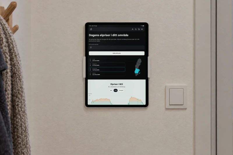 Tibber's page Dagens elpriser i ditt område, today's electricity prices in Sweden's four price areas, on a tablet mounted on the wall of a hallway beside a light switch, with a winter coat on a hook and snow outside the door