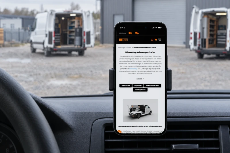 Work System's page Bilinredning Volkswagen Crafter on a phone in a holder on the dashboard of a work van, with two white vans with their rear doors open in the workshop yard outside the windscreen