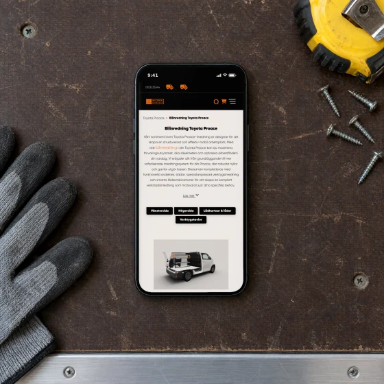 Work System's page “Bilinredning Toyota Proace” on a phone lying on the plywood load floor of a work van, beside a pair of work gloves, a tape measure and a few loose screws.